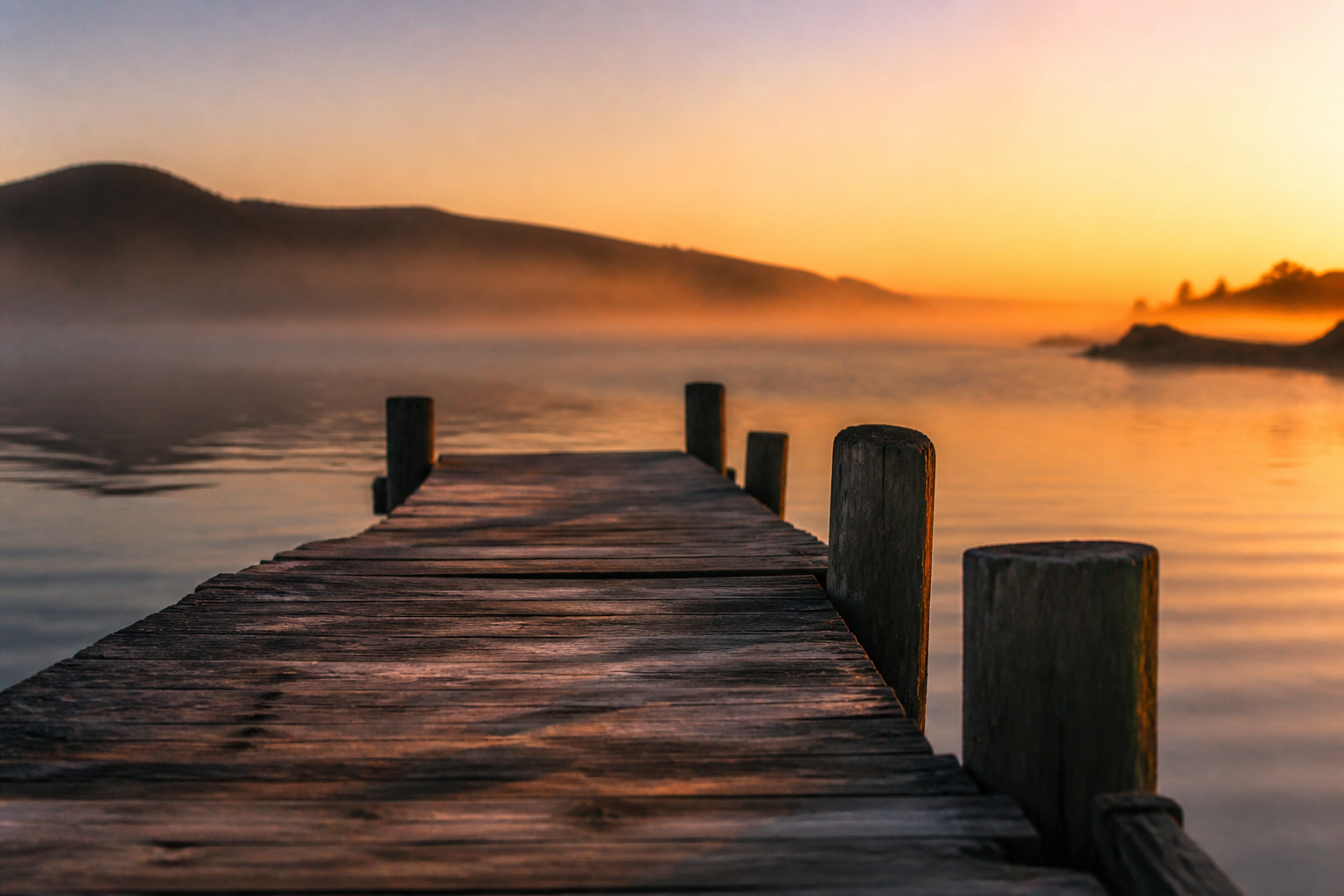 A weathered wooden dock stretching into still water at golden hour, with mist over the bay and distant hills.